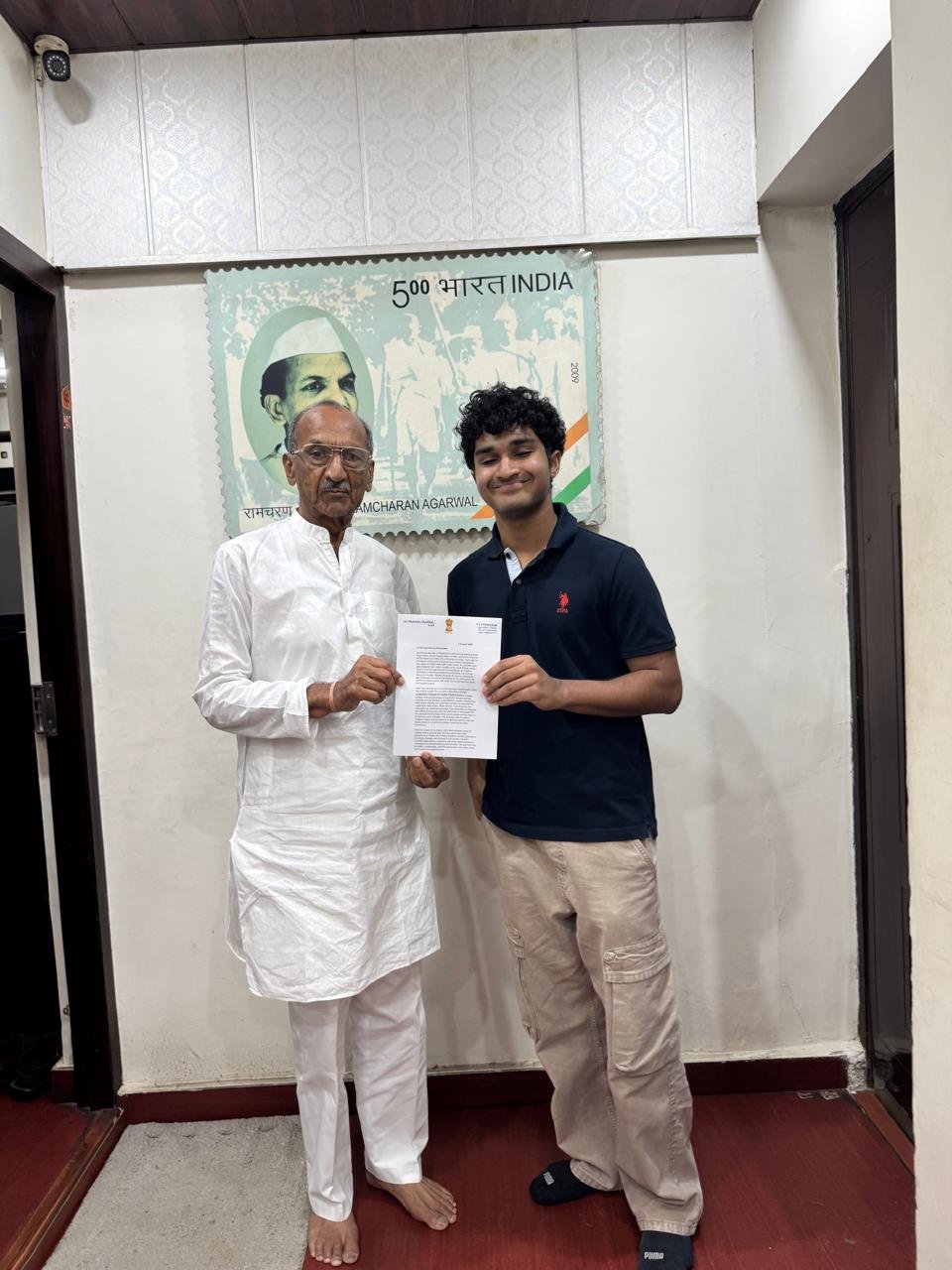 Standing with Shri Jai Prakash Agarwal, holding the signed accessibility petition, beneath a commemorative postage stamp of Shri Ramcharan Agarwal