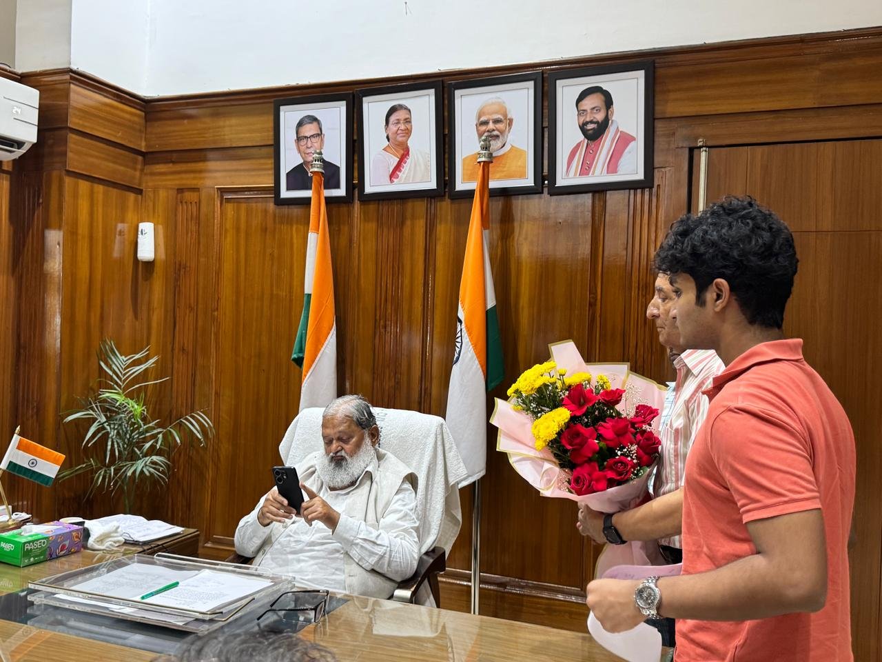 Approaching Anil Vij's desk with a bouquet of flowers as he speaks with an aide