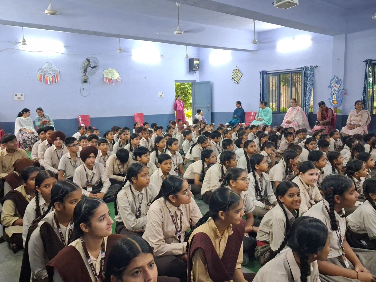 A wider view of the school assembly, with teachers seated along the back wall as students listen